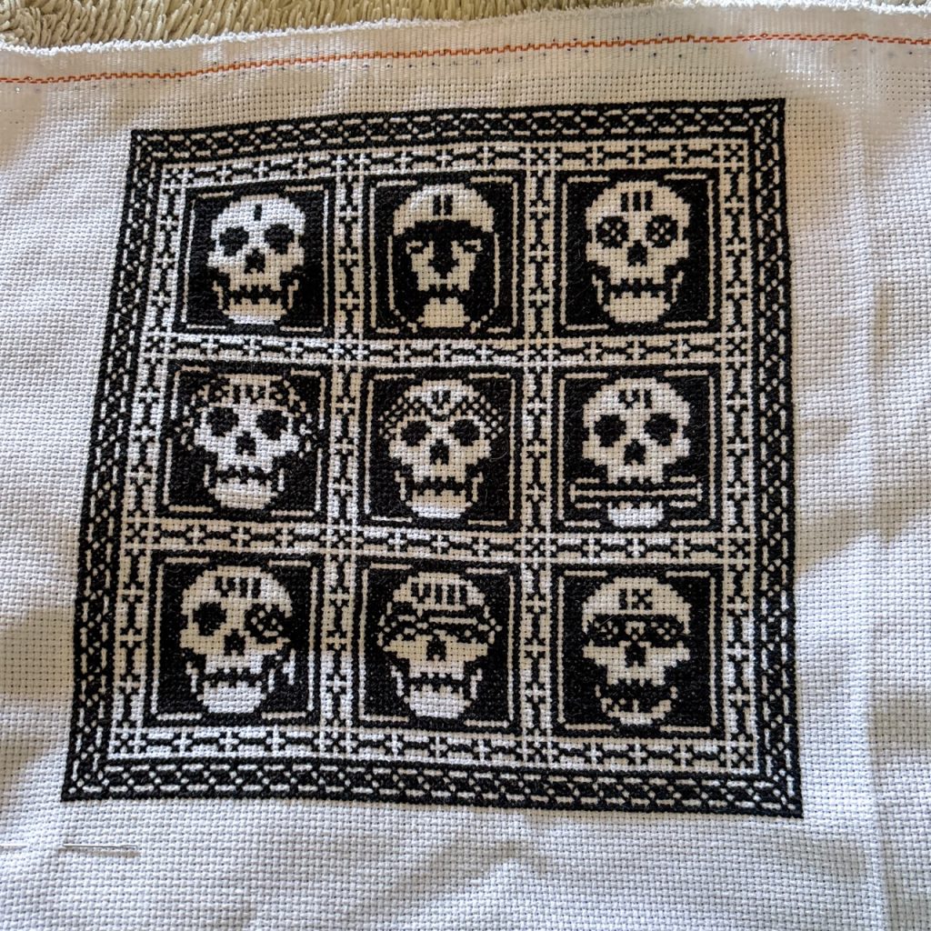 A slightly rumpled cross stitch, all black on white aida, depicting 9 skulls that represent the 9 houses in Gideon the Ninth by Tamsyn Muir.